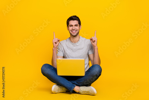 Full length photo of funny brunet millennial guy sit with laptop index up wear t-shirt jeans shoes isolated on yellow background