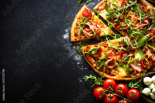 Pizza on black background. Traditional italian pizza with ham, cheese, tomatoes and arugula. Flat lay with space for text.