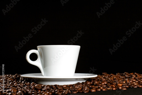 Hot coffee in a white coffee cup and many coffee beans placed around on dark background, with copy space.
