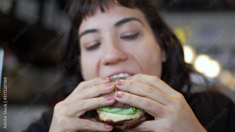 Closeup hand holding hamburguer. Young woman eating burger at restaurant. Girl takes a bite of fast food Closeup hand holding hamburguer. Young woman eating burger at restaurant. Girl takes a bite of fast food