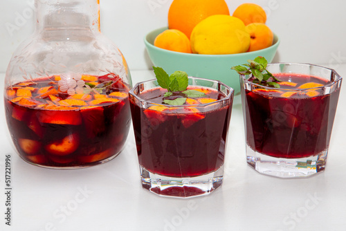 Full jar and two glasses of homemade red wine sangria cocktails ice and mint inside, with a bowl of orange, lemon, apricot background on a white table. copyspace