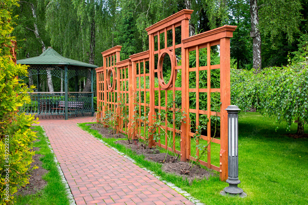 Vertical wooden pergola with square pattern in growth roses garden with stone brick way and ground lantern in backyard with different trees and lawn with iron forged gazebo on background, nobody. Vertical wooden pergola with square pattern in growth roses garden with stone brick way and ground lantern in backyard with different trees and lawn with iron forged gazebo on background, nobody.