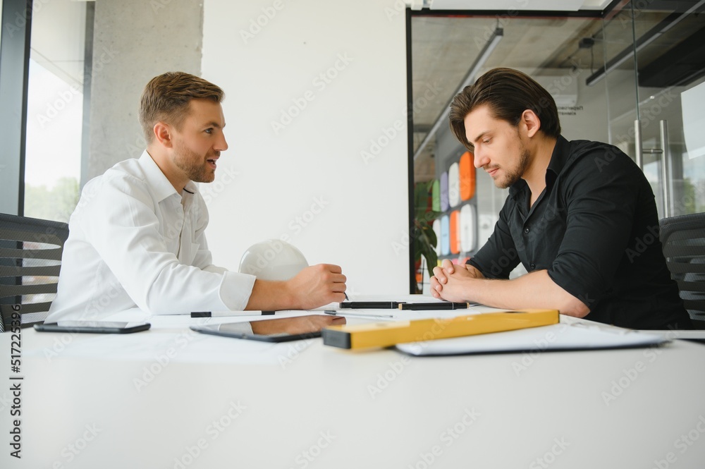 Two colleagues discussing data working on architectural project at construction site at desk in office Two colleagues discussing data working on architectural project at construction site at desk in office