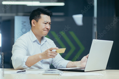 Worried young Asian businessman looking surprised at laptop and credit card in hand. Sitting in the office at the desk