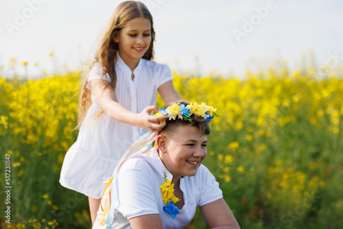 Sister braids ribbons in Ukrainian wreath with flowers on older brother s head  on meadow against field