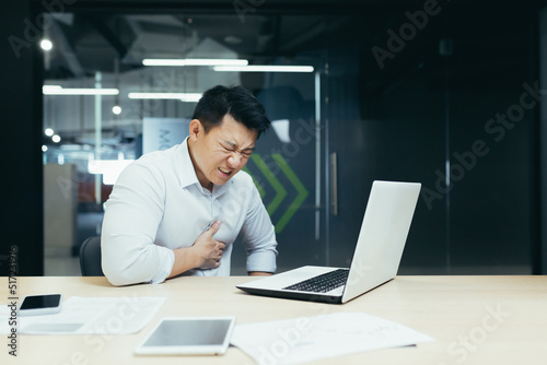 Heart attack at work. A young Asian man, businessman, office worker holds his heart, feels pain, had a stroke, heart attack at the desk in the office.