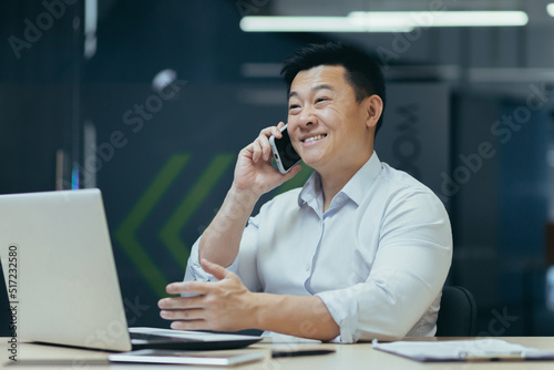 Online store. A young Asian salesman talks on the phone with customers, offers them to buy goods. Sitting behind a desk in a modern office. © Liubomir