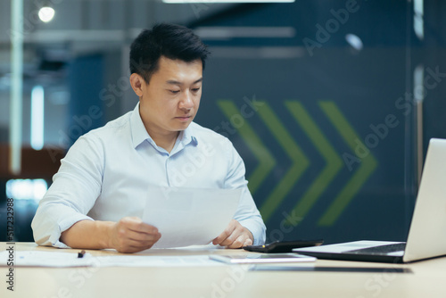 Portrait of young serious and focused Asian male accountant working with documents and calculator. Sitting at a desk with a laptop in a modern office