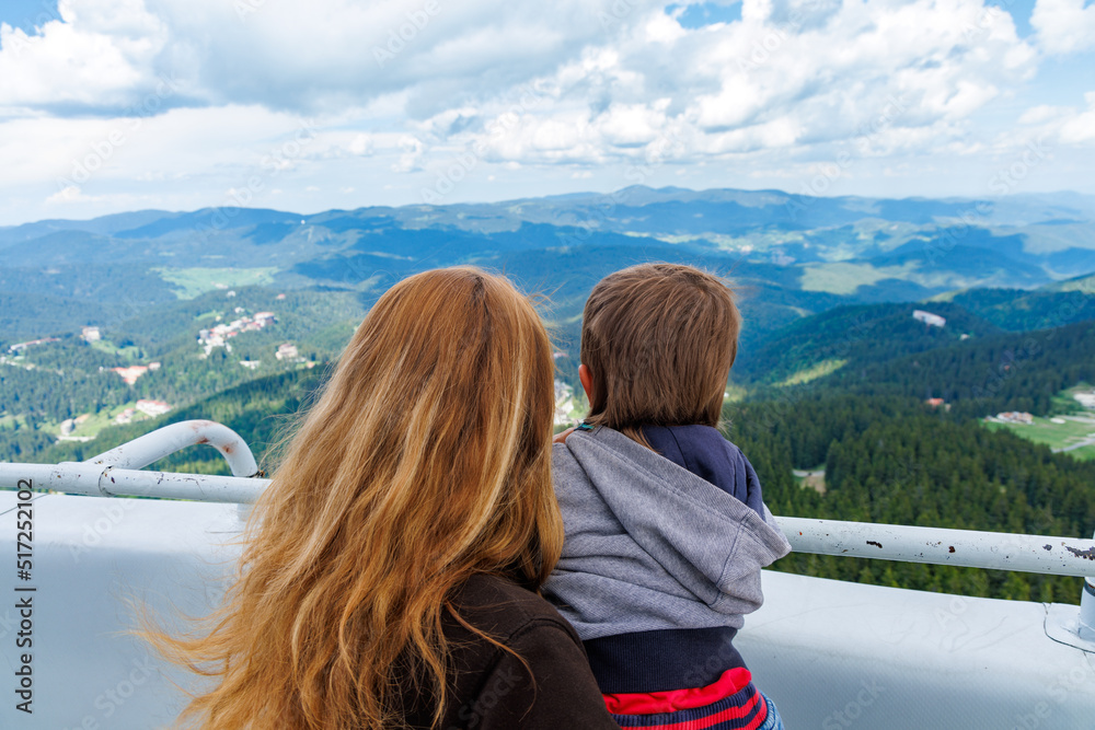 Fototapeta premium Mother holds son and shows him landscapes of forest valley of Rhodope Mountains while standing on tower of Snezhan