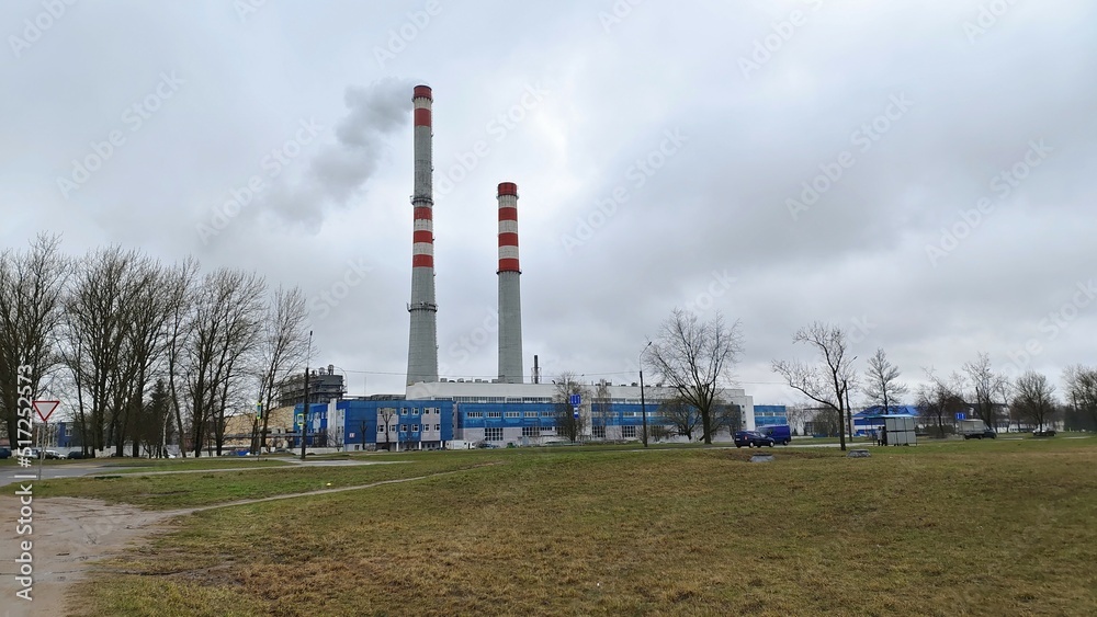 The factory stands on the outskirts of the city by the paved road on which cars move. Smoke is coming out of the plant's pipes. There is a grassy lawn in front of the plant, where trees grow. Cloudy