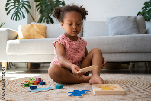 African american cute girl is playing educational logic games. Child with wooden puzzle toys. Beautiful baby girl playing at home alone with her children's coloured wooden toys for development