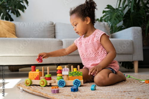 Educational eco-friendly toys. Afro two years old child girl playing with wooden colourful train at home. Development of kids fine motor skills, imagination and logical thinking concept