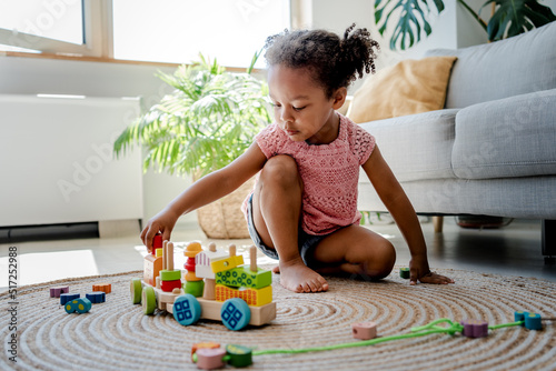 Excited little black girl have fun play toys on warm floor at living room. Young female afro baby engaged in education game with wooden railway station