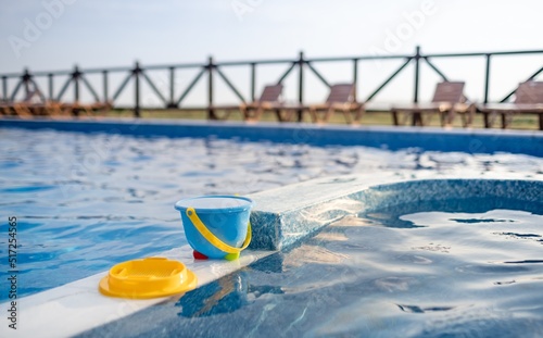 A children's bucket with sieve of water and some toys are on the boat of pool against the background of summer sunset