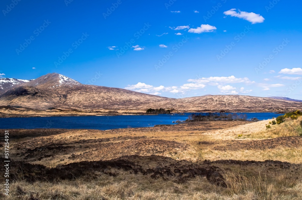 Beautiful calm loch or lake surrounded by rugged hills, brown scrub grassland and blue sky with a few clouds. Copy space. Scottish Highlands, Scotland.