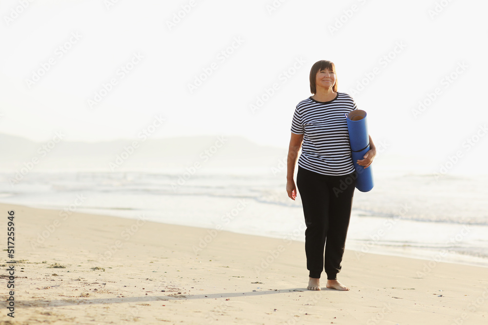 Middle age woman holding a sports mat and preparing to practice yoga outdoors on sea beach. Happy mature overweight woman exercising on seashore. copy space. Meditation, yoga and relaxation concept.