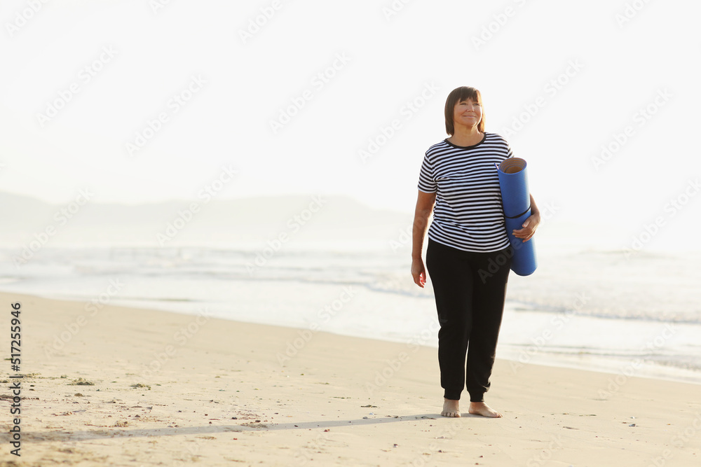 Middle age woman holding a sports mat and preparing to practice yoga outdoors on sea beach. Happy mature overweight woman exercising on seashore. copy space. Meditation, yoga and relaxation concept. Middle age woman holding a sports mat and preparing to practice yoga outdoors on sea beach. Happy mature overweight woman exercising on seashore. copy space. Meditation, yoga and relaxation concept.