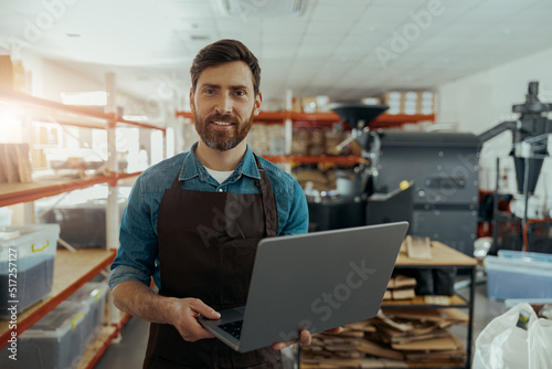 Smiling male worker in uniform standing on coffee warehouse with laptop