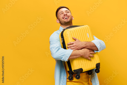 Fototapeta Enjoyed funny smiling tanned handsome man in blue shirt hugs hold suitcase ready for vacation posing isolated on orange yellow studio background