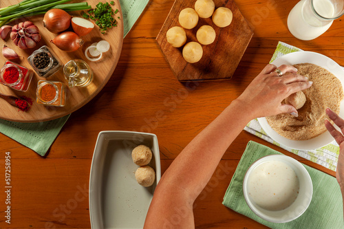 Woman hands preparing brazilian croquette (coxinha de frango) with breadcrumbs on a wooden kitchen table - Top view