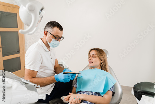 Woman looking at veneers or implants teeth color matching samples in doctor hands. Dentistry. Dentist demonstrate teeth color shades guide for tooth whitening for woman patient in dental clinic.