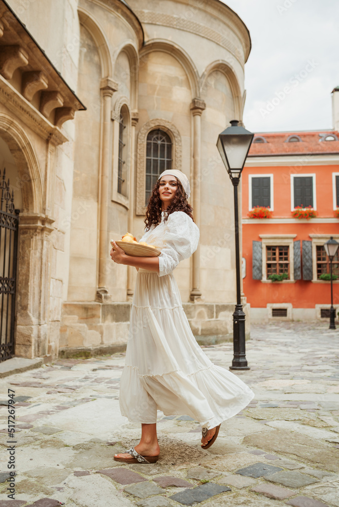 Beautiful curly brunette woman wearing trendy summer white linen shirt, maxi skirt, holding bowl with lemons, walking in sunny street of European city. Full-length outdoor portrait