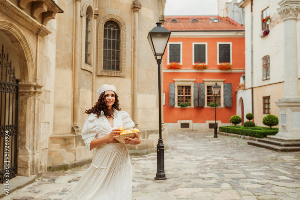 Happy smiling curly brunette woman wearing trendy summer white linen shirt, long skirt, bandana, holding bowl with lemons, posing in street of European city. Copy, empty space for text