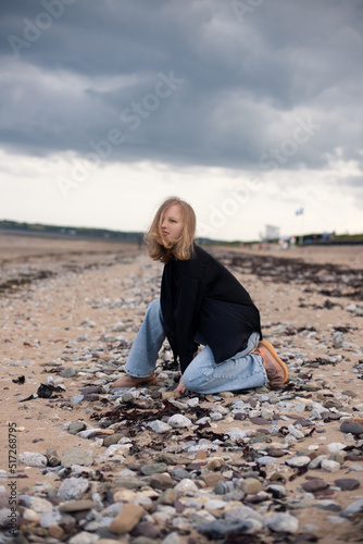 Young blonde girl dressed in a black silk shirt and blue jeans is walking along the beach and enjoying the cloudy weather