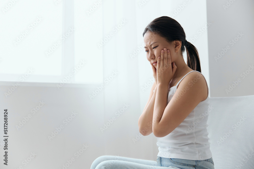 Caries Dental decay Wisdom teeth Pulpitis Periodontitis. Unhappy suffering tanned beautiful young Asian woman touch cheek at home interior living room. Injuries Poor health Illness concept. Cool offer Caries Dental decay Wisdom teeth Pulpitis Periodontitis. Unhappy suffering tanned beautiful young Asian woman touch cheek at home interior living room. Injuries Poor health Illness concept. Cool offer