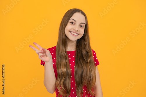 Cheaky teen girl giving victory sign. Happy teenager child smiling with v-sign, positive gesture, standing against yellow isolated background.