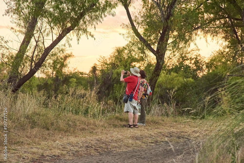 Fototapeta premium Couple of mature travelers taking pictures and enjoying the scenery at sunset at the Multiple Uses Natural Area, River of the Birds Park (Parque Rio de los Pájaros) in Colon, Entre Rios, Argentina.