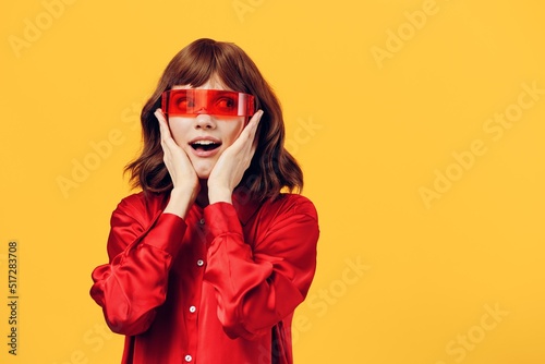  happy, stylish woman stands on a yellow background in a red shirt and fashionable glasses of an interesting shape, smiling happily with her hands near her face. Horizontal photo with empty space