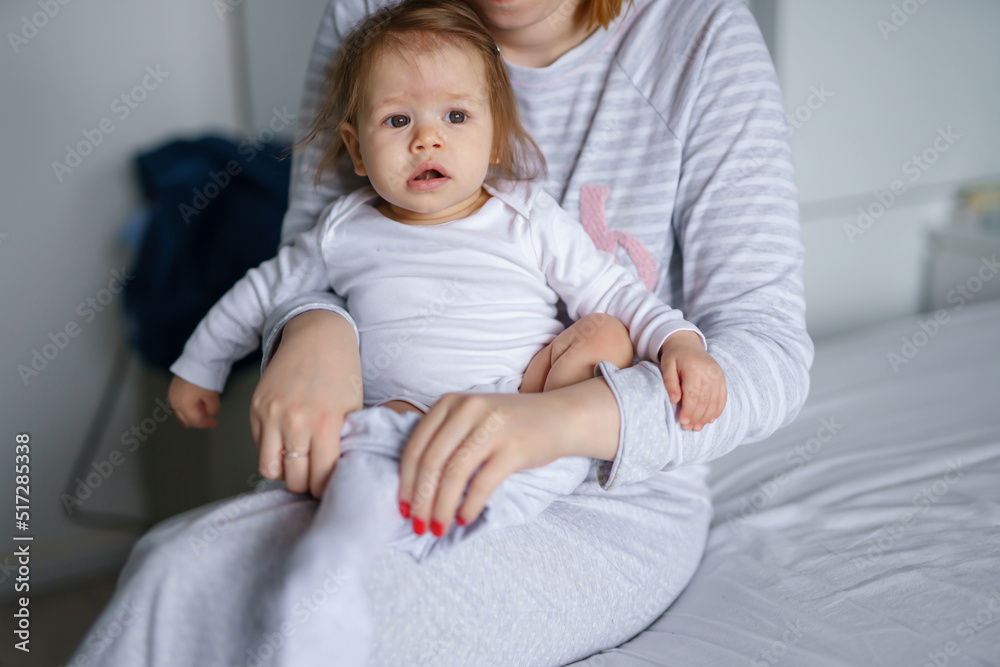 One caucasian baby sitting on the bed in bright room in day while her mother is changing her clothes getting dressed real people care growing up and family concept copy space One caucasian baby sitting on the bed in bright room in day while her mother is changing her clothes getting dressed real people care growing up and family concept copy space