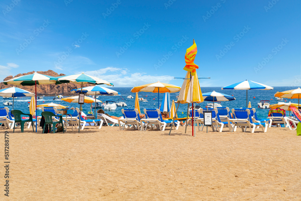 Beach chairs and colorful umbrellas along the sandy Platja Gran beach at the resort town of Tossa de Mar, Spain, on the Costa Brava coast of the Mediterranean Sea.