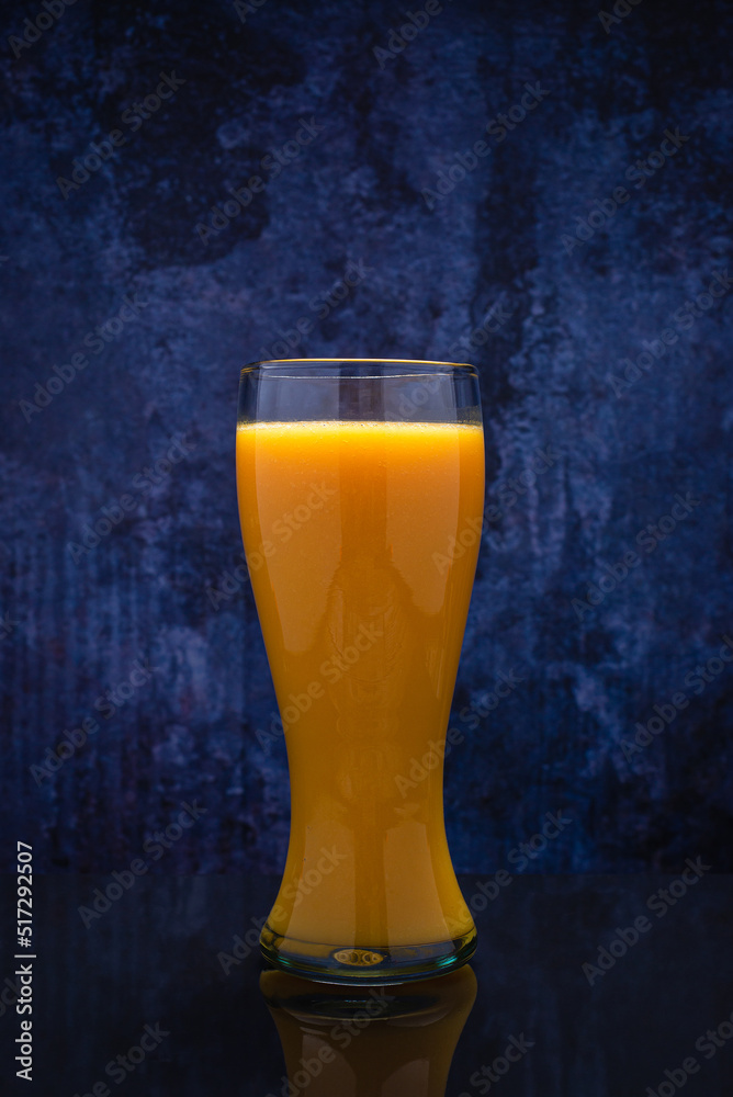 Tall glass with mango juice on black glass table with reflection. Blue background.