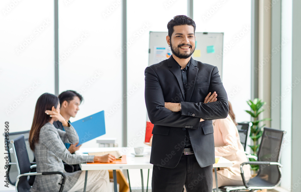 Portrait shot of millennial Indian Asian professional successful bearded businessman in formal suit standing posing smiling crossed arms in meeting room while employee and colleague brainstorming Portrait shot of millennial Indian Asian professional successful bearded businessman in formal suit standing posing smiling crossed arms in meeting room while employee and colleague brainstorming