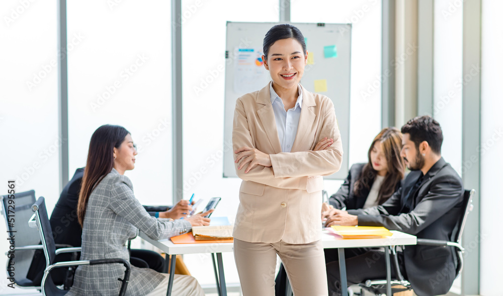 Obraz premium Portrait shot of millennial Asian cheerful successful professional businesswoman entrepreneur in formal suit standing smiling crossed arms in meeting room while employee and colleague brainstorming