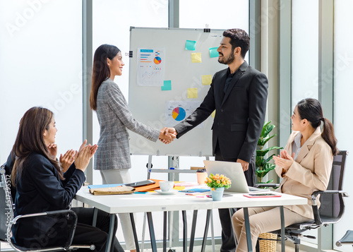 Asian professional businesswoman and Indian male successful businessman in formal suit standing shaking hands when business agreement deal done while female colleagues clapping hands celebrating