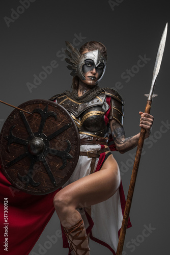 Shot of female warrior from past with shield and spear dressed in armor and red cape looking at camera.