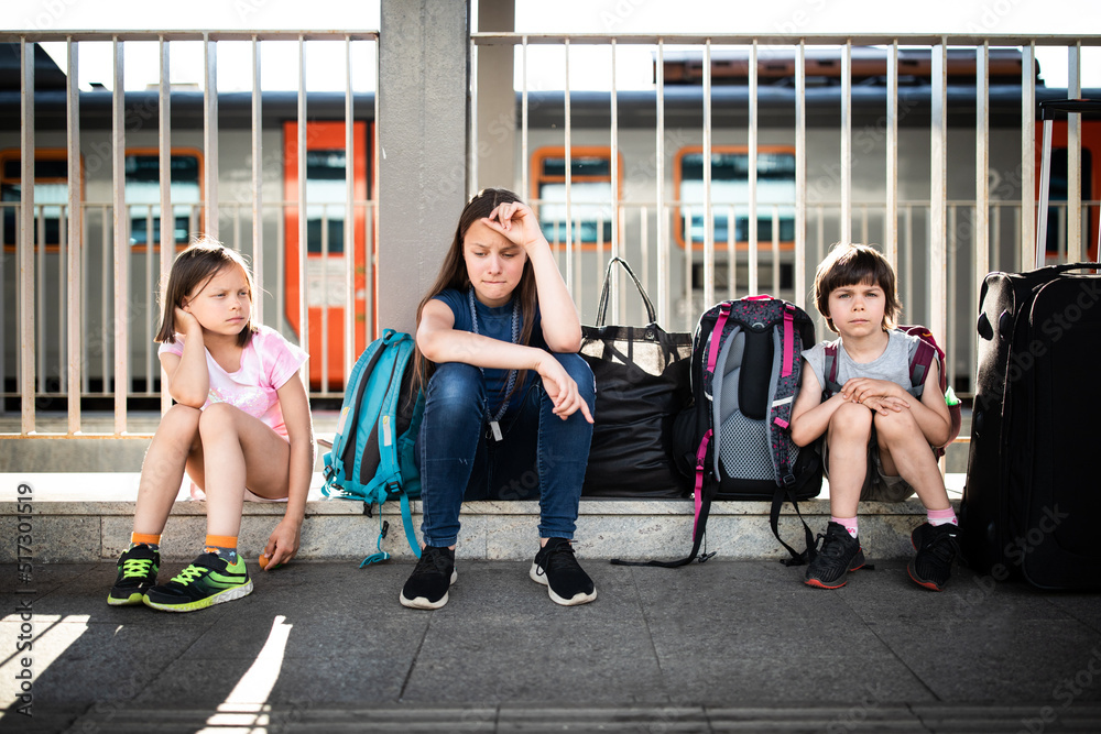 Three sad children sitting on platform on railway station with luggage and waiting for train on sunny summer day