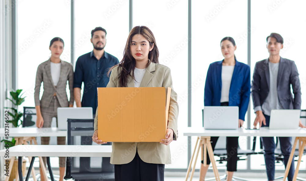 Portrait shot of Asian sad jobless businesswoman in casual suit standing holding belongings in cardboard box after fired while male and female colleagues waving hands goodbye in blurred background