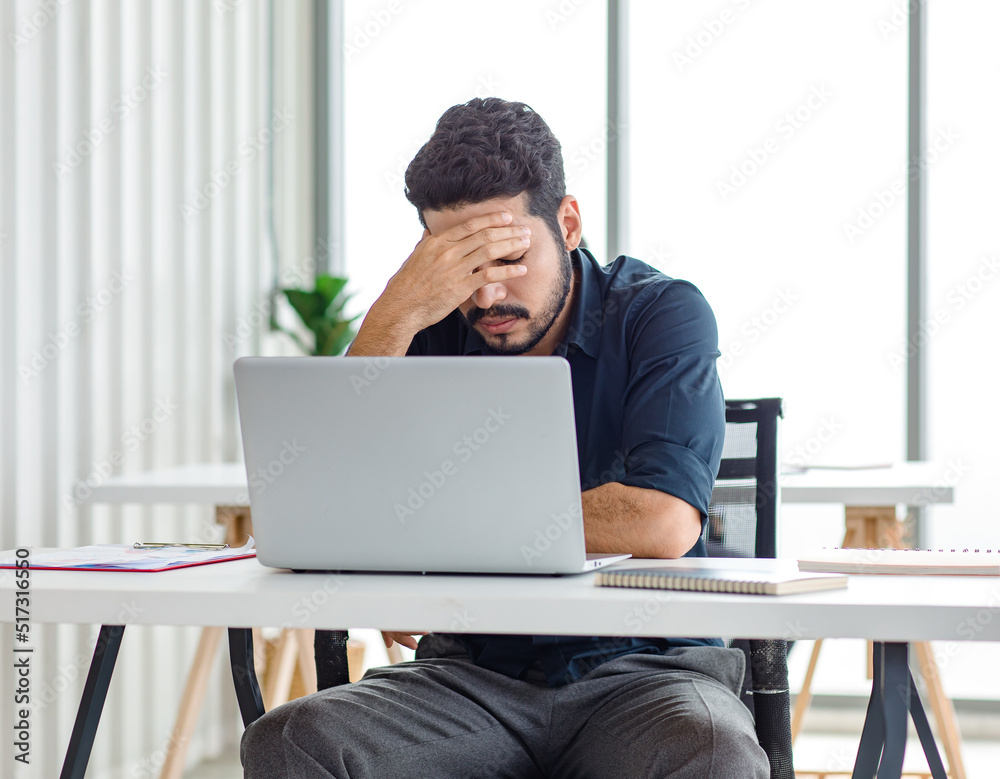 Indian stressed depressed exhausted bearded male businessman sitting at workstation with laptop computer close eyes hold hand on head feeling tired and sleepy after working late overtime last night Indian stressed depressed exhausted bearded male businessman sitting at workstation with laptop computer close eyes hold hand on head feeling tired and sleepy after working late overtime last night
