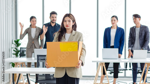 Portrait shot of Asian sad jobless businesswoman in casual suit standing holding belongings in cardboard box after fired while male and female colleagues waving hands goodbye in blurred background