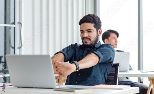 Indian millennial tired exhausted sleepy bearded dull face male businessman sitting at workstation desk with laptop computer stretching arms widely in morning after working late overtime last night