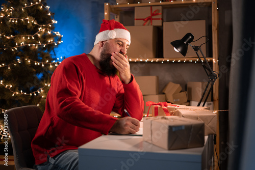 Small and medium business start up concept Young aspiring businessman hindu man, small business owner in santa claus costume tired working at night in home office. Yawns at the table. © mtrlin