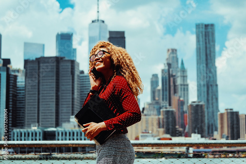 Portrait smiling young adult entrepreneur millennial woman with eyeglasses and afro hair talking on a phone call outdoors with Manhattan New York City skyline skyscraper behind Hudson river