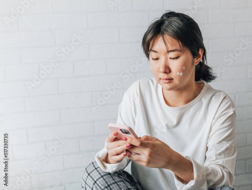 girl is texting on a smartphone while sitting on a bed at home. copy space. teenage girl with problematic skin, with a therapeutic cream applied.