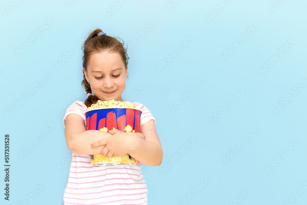 little girl joyfully presses a huge glass of popcorn to herself with both hands and looks at him tenderly, on a blue background, copy space little girl joyfully presses a huge glass of popcorn to herself with both hands and looks at him tenderly, on a blue background, copy space