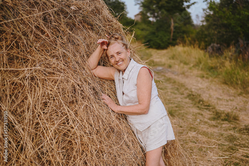 Elderly senior traveling backpacker mature woman tourist smiling posing with stack of hay in field outdoors in countryside. Retired people summer holiday vacation, active lifestyle concept