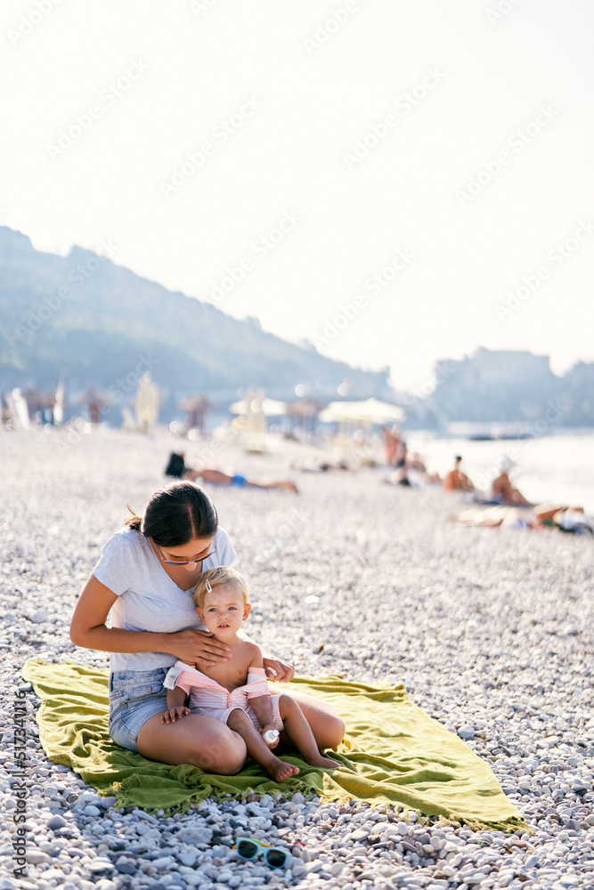 Mom applying sunscreen to a little girl on a blanket on the beach